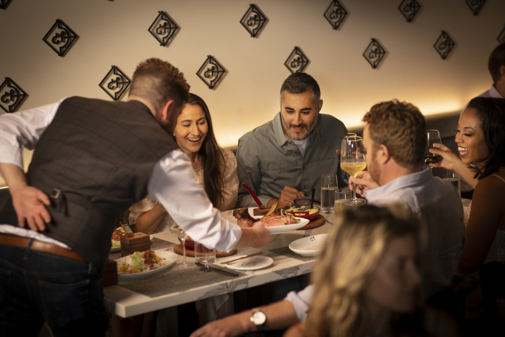 Group Of People Enjoying Prime Steak Being Presented By A Server At Huntress